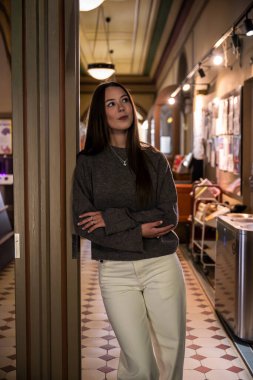 Gri kazaklı ve pantolonlu genç bir kadın klasik koridorda duruyor.