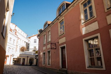 Eski Tallinn kasabasının arka planında mavi bir gökyüzü ve antik binaların yanına park edilmiş modern arabaların olduğu bir cadde manzarası.