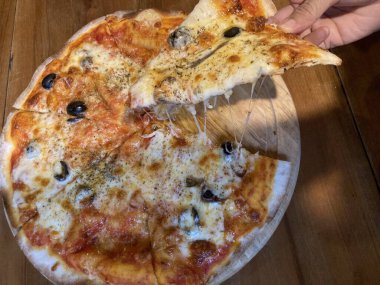 Sıcak güneş ışığı dairesel ahşap pizza tepsisinde parlarken bir kadının eli Napoli pizzasından bir dilim kapıyor.