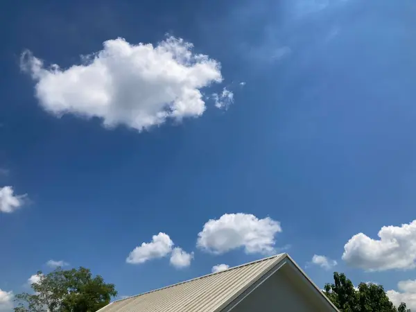 Evin beyaz çatısının üst manzarası, süper parlak mavi gökyüzü ve evin yanında yeşil ağaçlarla süzülen tüylü beyaz bulutlar.