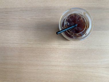 Açık plastik bir fincanda buzlu sade kahve ve siyah plastik bir pipet açık kahverengi bir ahşap masada güzel ahşap tahıllar ve çerçevenin sol tarafında fotokopi alanı ile oturmaktadır.