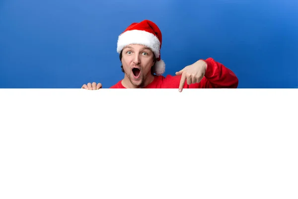驚きの男の写真は 赤い服とXマスの帽子を着用青い色の背景に隔離された空のスペースポスター広告を保持し クリスマス 新年のコンセプト — ストック写真