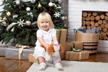 Beyaz elbiseli çok hoş bir sarışın kız hediye kutusunda oturuyor ve evin parlak iç kısımlarındaki Noel ağaçlarının arka planında yüksek sesle gülüyor. Tatiller ve çocukluk kavramı 