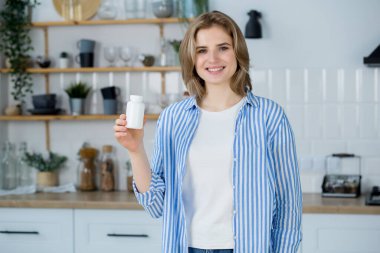 Mutfakta elinde besin takviyeleri şişesiyle gülümseyen beyaz bir kadın kameraya bakıyordu. Sağlıklı konsept