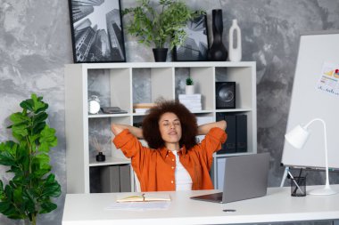 Mutlu, rahat, Afro-Amerikan genç kadın bilgisayar işinden kafası karışmış bir şekilde koltukta dinleniyor, negatif duyguları rahatlatıyor. Kadın işyerinde rahat otur kestir ya da hayal kur.