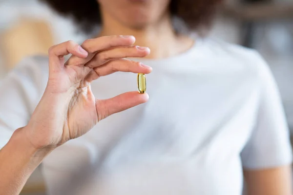 African american woman holds in her hand a yellow transparent capsule with vitamin D or omega-3 close-up. Preventive medicine. Woman holding fish oil capsule.