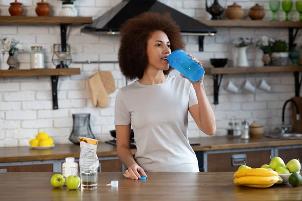 Portrait of fit black woman in sportswear at the kitchen is drinking from shaker with whey protein or water. Healthy sporty lifestyle