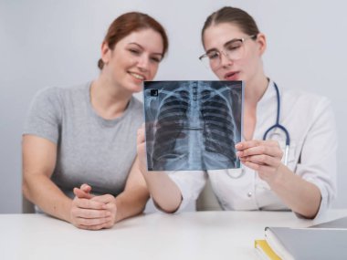 Doktor beyaz bir kadına danışıyor ve akciğerlerin röntgenleri hakkında yorum yapıyor.