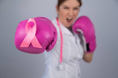 Pembe boks eldivenli, gri arka planda pembe kurdeleli bir kadın doktor. Göğüs kanserine karşı savaş