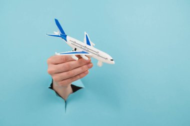 Mavi arka planda bir delikten dışarı sarkan bir kadın el bir uçak modeli tutar.
