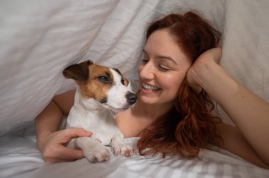 Kafkasyalı kızıl saçlı kadın beyaz çarşafta Jack Russell teriyer köpeğiyle kucak kucağa uyuyor.