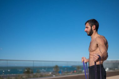 Tişörtsüz adam dışarıda lastik bantlarla egzersiz yapıyor.