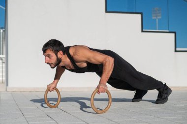 Siyah spor kıyafetli bir adam dışarıda ring şınavı çekiyor.