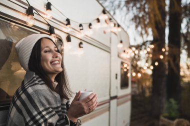 Ekoseye sarılı örgü şapkalı beyaz bir kadın dışarıda sıcak bir içecek içiyor. Sonbaharda bir karavanla seyahat et.