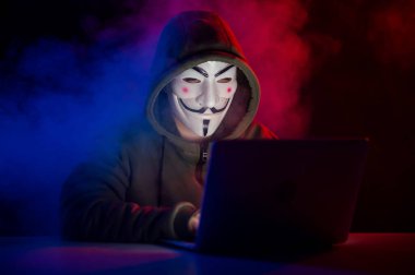5 Haziran 2022 Novosibirsk, Rusya: Kapüşonlu Anonymous kırmızı-mavi dumanla karanlıkta dizüstü bilgisayarda yazıyor