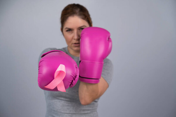Caucasian woman in pink boxing gloves with a pink ribbon on her chest on a gray background. Fight against breast cancer