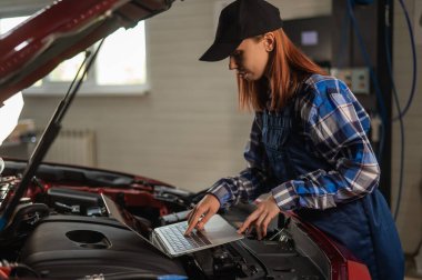 Kadın oto tamircisi dizüstü bilgisayar kullanarak motor kontrolü yapıyor.