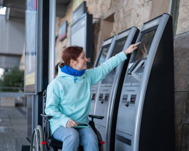 Tekerlekli sandalyedeki beyaz kadın self-servis kasasından tren bileti alıyor.