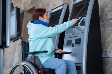 Tekerlekli sandalyedeki beyaz bir kadın, bir self-servis kasasında cep telefonu kullanarak tren bileti alıyor.
