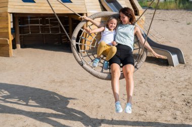 Anne ve kızı salıncakta sallanıyor. Beyaz kadın ve küçük kız oyun alanında eğleniyor.