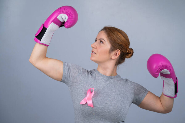 A woman with a pink ribbon on her chest raises her hands in pink boxing gloves on a gray background. Victory over breast cancer