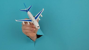 Mavi arka planda bir delikten dışarı sarkan bir kadın el bir uçak modeli tutar.