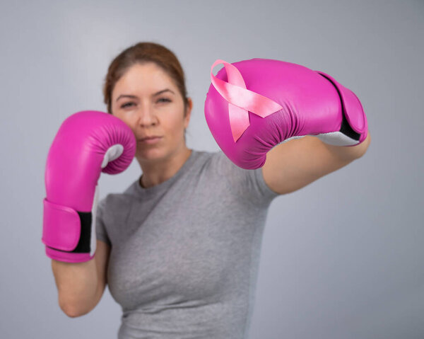 Caucasian woman in pink boxing gloves with a pink ribbon on her chest on a gray background. Fight against breast cancer