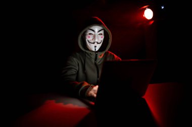 5 Haziran 2022 Novosibirsk, Rusya: Kapüşonlu Anonymous kırmızı ışıkta karanlıkta dizüstü bilgisayarda yazıyor
