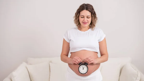 裸の腹を持つ妊婦は目覚まし時計を持っている 出生の予定日 — ストック写真