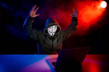 5 Haziran 2022 Novosibirsk, Rusya: Kapüşonlu Anonymous kırmızı-mavi dumanla karanlıkta dizüstü bilgisayarda yazıyor