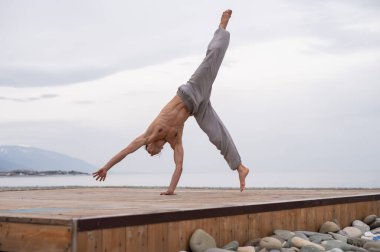 Üstsüz beyaz adam sahilde akrobatik tekerlek yapıyor.