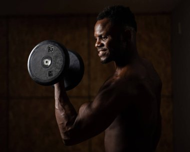 Çekici Afrikalı Amerikalı adam gülümsüyor ve halterlerle egzersiz yapıyor.