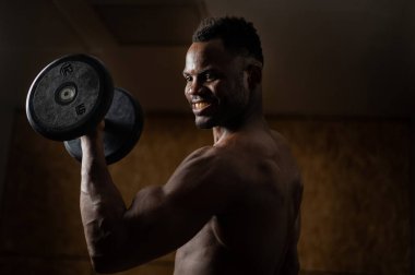 Çekici Afrikalı Amerikalı adam gülümsüyor ve halterlerle egzersiz yapıyor.