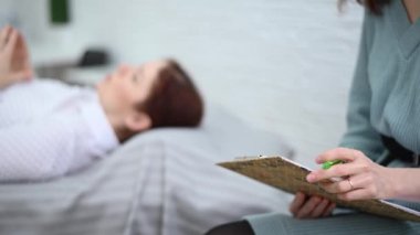 Bir kadın psikoterapist bir hastayla seans sırasında tablete not alır. Kafkasyalı kadın psikoloğun kanepesinde yatıyor.