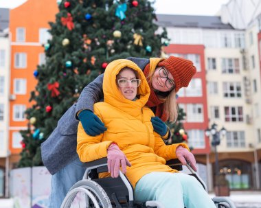 Kafkasyalı bir kadın arkadaşını Noel ağacının yanında tekerlekli sandalyeye oturtuyor.