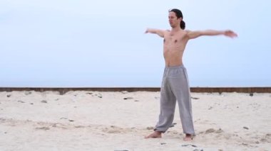 Beyaz bir adam, çıplak gövdesiyle deniz kıyısında wushu yapıyor.