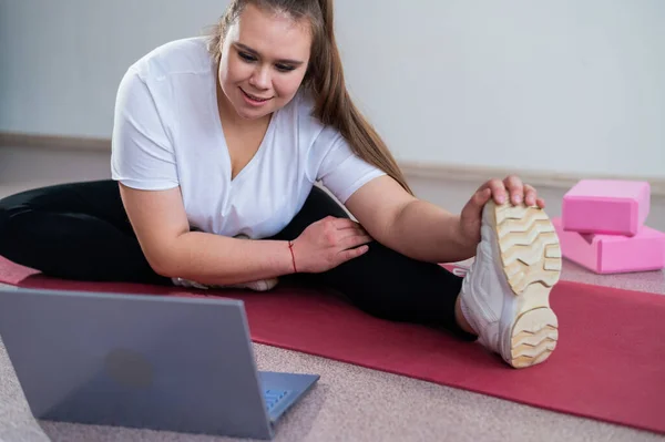 年轻的高加索胖女人弯腰躺在运动垫上 在笔记本电脑上看训练录像 一个胖胖的女孩在用视频通信进行远程伸展 — 图库照片