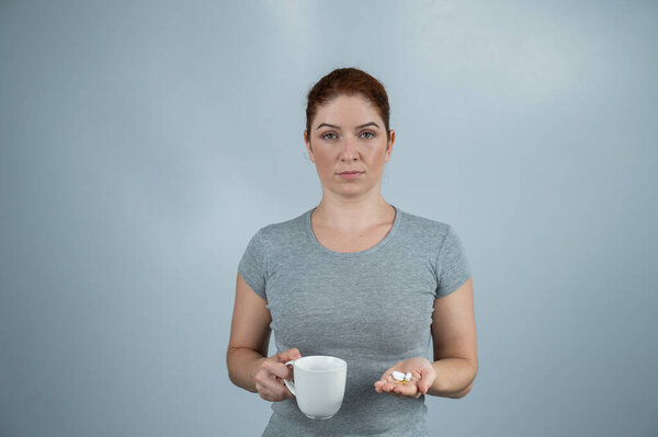 Caucasian woman holding handful of pills and mug on gray background