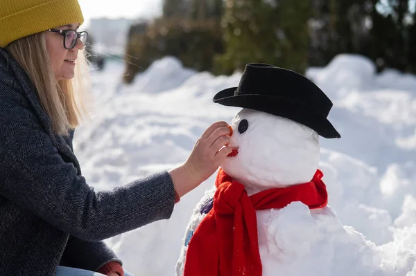 Caucasian woman sculpts a snowman from the snow. The carrot is the place of the nose