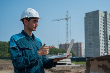 Kasklı bir erkek müteahhit elinde dizüstü bilgisayarla inşaat alanının arka planında duruyor.