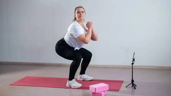 A chubby young woman is watching an online fitness lesson on a mobile phone. Distance sports training.