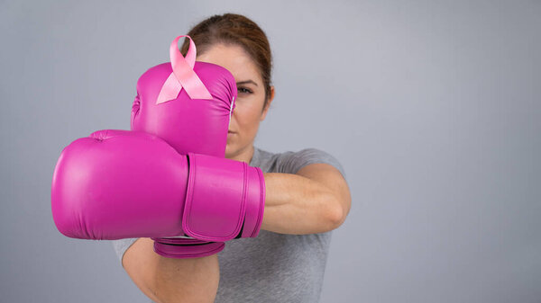 Caucasian woman in pink boxing gloves with a pink ribbon on her chest on a gray background. Fight against breast cancer