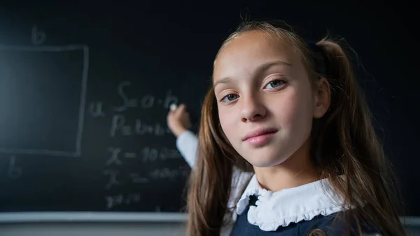 Portrait of a caucasian girl in the classroom. The schoolgirl writes the formula with chalk on the blackboard and looks into the camera