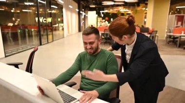 Beyaz kadın, erkek masasında duruyor. Patron, astının çalışmasını onaylıyor.