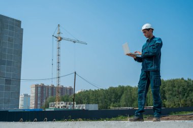 Kasklı bir erkek müteahhit elinde dizüstü bilgisayarla inşaat alanının arka planında duruyor.