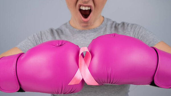 Caucasian woman in pink boxing gloves with a pink ribbon on her chest on a gray background. Fight against breast cancer