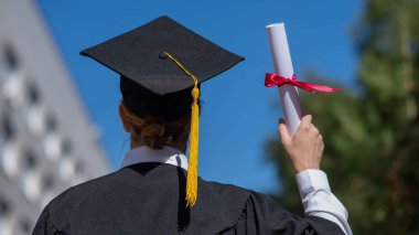 Mezuniyet elbisesi giymiş, elinde diplomasıyla üniversitenin yanında duran beyaz bir kadının arka görünüşü.