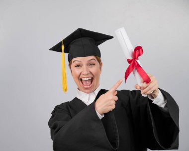 Mezuniyet elbisesi giymiş mutlu bir kadın diplomasını işaret ediyor.