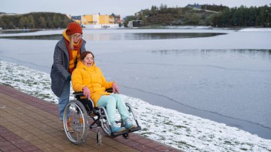Beyaz kadın korkmuş arkadaşını gölün kenarındaki tekerlekli sandalyeye yuvarlıyor.