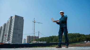 Kasklı bir erkek müteahhit elinde dizüstü bilgisayarla inşaat alanının arka planında duruyor.